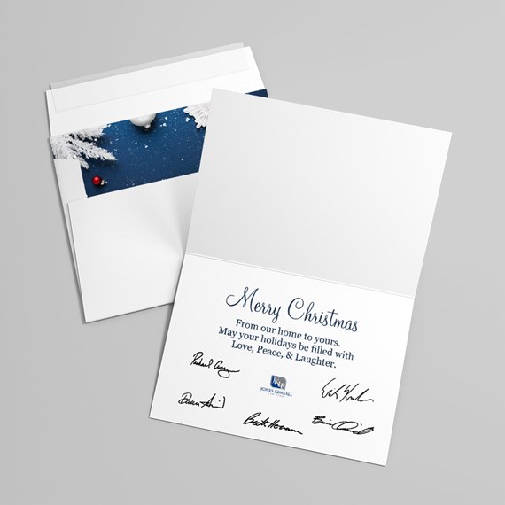 Folded white Christmas card with navy 'Merry Christmas' greeting and printed signatures, shown with an open envelope featuring a dark blue snowy liner and white frosted pine with a red ornament