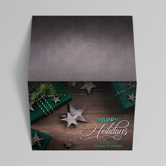 Folded holiday card with green gift boxes, gray star ornaments, pine sprigs and twine on a wood background