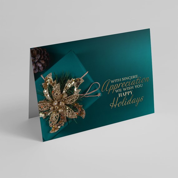 Folded teal holiday card with gold foil script and a glittery gold poinsettia ornament with pine sprigs