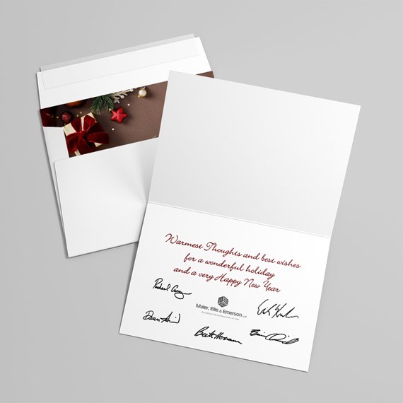 White folded holiday greeting card open to show red cursive message and signatures, with matching envelope and decorative insert featuring a wrapped gift and ornaments
