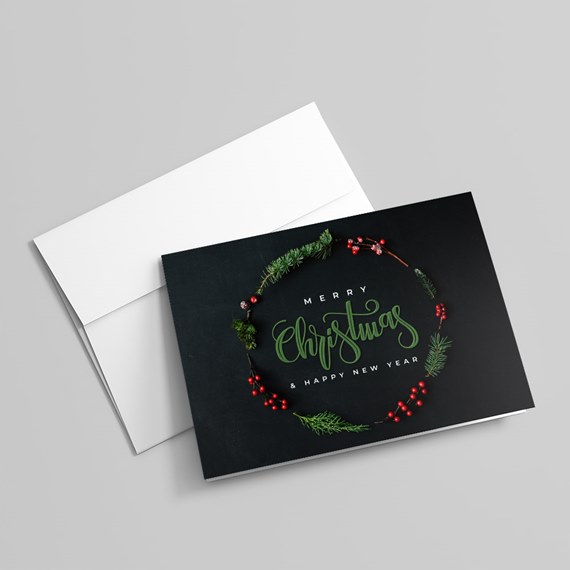 Black Christmas greeting card with green script and pine wreath with red berries, accompanied by a white envelope