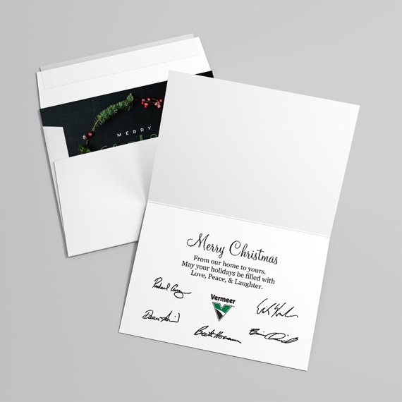 White folded Christmas greeting card open to show printed message, signatures and logo, with envelope and wreath-themed black insert visible.