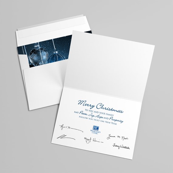 Folded white Christmas card with blue "Merry Christmas" text and signatures, next to a white envelope with a blue ornament liner