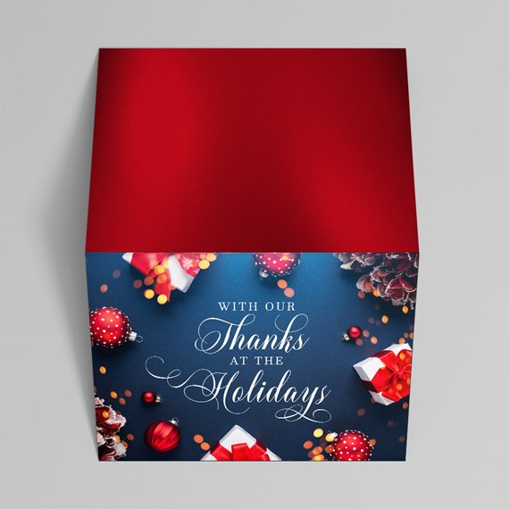 Folded holiday greeting card with red flap, navy front, white script text, red ornaments and gift boxes with red bows.