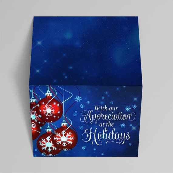 Folded holiday greeting card with blue starry background, red hanging ornaments with white snowflakes and white script greeting