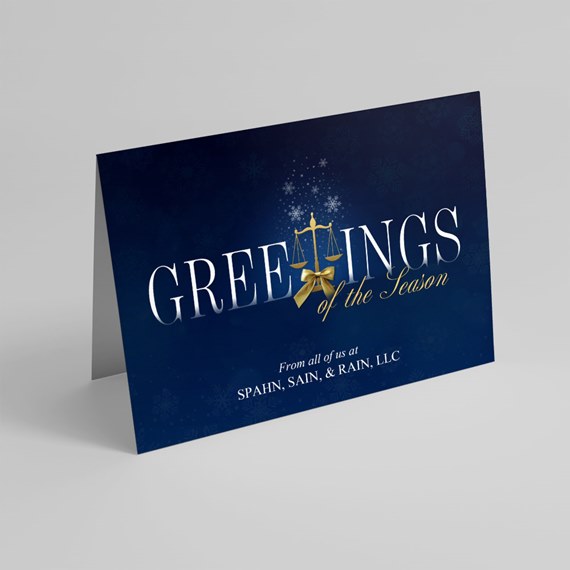 folded navy holiday card with white serif text, gold scales and bow, and snowflake motif